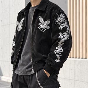 Peut inclure: Une veste noire zippée avec des broderies blanches représentant un aigle sur la poitrine et un long dragon sinueux le long de la manche. La veste présente un col, des poignets côtelés et un t-shirt gris foncé porté en dessous.