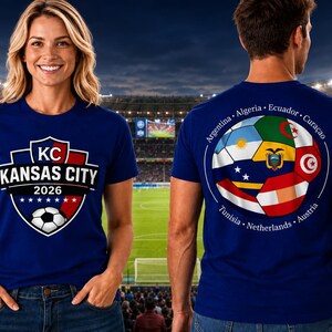 May include: A royal blue short-sleeve t-shirt. The front features a shield graphic with the text KC KANSAS CITY 2026 and a soccer ball. The back displays a circular graphic of a soccer ball composed of various national flags and country names.