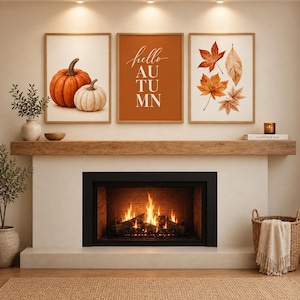 May include: Three framed wall art prints displayed above a fireplace. The left print shows two pumpkins, the center print features the text "hello AUTUMN" on an orange background, and the right print displays a collection of various autumn leaves.