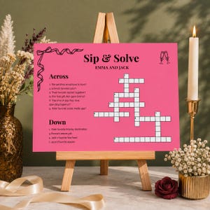 Könnte beinhalten: Ein leuchtend rosa rechteckiges Schild mit der Überschrift Sip & Solve Emma and Jack, das ein Kreuzworträtsel mit nummerierten Hinweisen für waagerecht und senkrecht enthält. Das Schild steht auf einer Holzstaffelei mit schwarzem Text und Grafiken.