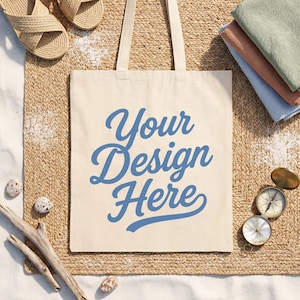 May include: A beige canvas tote bag with the blue text Your Design Here printed on the front. The bag features two long shoulder straps and is displayed alongside a pair of woven sandals, a stack of folded fabric, and a brass compass.