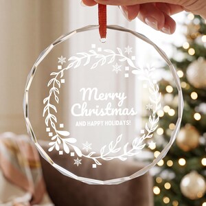 May include: A circular, faceted clear glass ornament featuring a white etched design of a leafy wreath, snowflakes, and the text Merry Christmas AND HAPPY HOLIDAYS! The ornament is suspended by a thin red ribbon for hanging purposes.