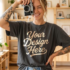 May include: A charcoal gray short-sleeve t-shirt featuring the text Your Design Here in a metallic gold script font. The shirt is worn with light blue denim jeans. The garment has a crew neckline and a relaxed fit with visible stitching.