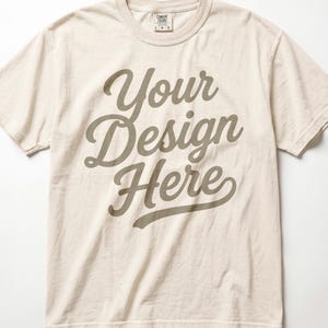 May include: A light beige short-sleeve t-shirt featuring the dark brown text Your Design Here printed in a cursive script font across the chest. The shirt has a crew neckline and a Comfort Colors brand label visible at the inner neck area.