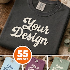 Puede incluir: Una camiseta de cuello redondo gris carbón con el texto blanco Your Design impreso en una fuente cursiva. La camiseta tiene una etiqueta de la marca Comfort Colors en el cuello. Se muestran tres camisetas adicionales en azul claro, morado y verde salvia.