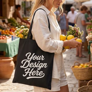 May include: A black canvas tote bag featuring the white text Your Design Here in a cursive script font. The bag has two shoulder straps and is shown in a market setting with yellow lemons and flowers tucked inside the main compartment.