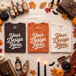 May include: Three t-shirts in dark brown, orange, and cream, each featuring the white text Your Design Here. The shirts are arranged with glass potion bottles labeled Witches Brew, Spider Venom, and Autumn Elixir, black candles, and pumpkins.