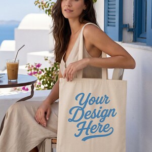 May include: A beige canvas tote bag featuring the blue text Your Design Here printed in a cursive script font on the front. The bag has two shoulder straps and a rectangular shape, presented as a blank mockup for custom graphic placement.