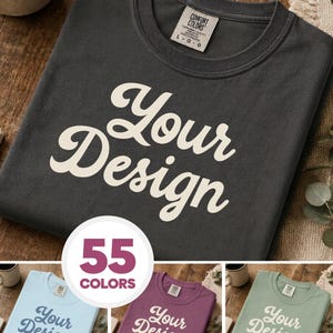 May include: A charcoal gray crewneck t-shirt featuring the text Your Design in a cream-colored script font. The shirt includes a Comfort Colors brand label on the inside collar. Three additional t-shirts in light blue, plum, and sage green are shown.