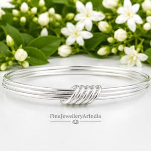 Op de afbeelding: Een gepolijste zilveren armband bestaande uit drie dunne, gladde ringen die in het midden zijn samengevoegd door een reeks van vijf kleine, strak gewikkelde zilveren draadspiralen. De tekst FineJewelleryArIndia staat onder de armband.