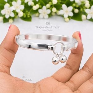 May include: A polished silver bangle bracelet featuring a circular center link with two small, round, dangling silver bells. The bracelet displays the text FineJewelleryArIndia in two locations, with a small heart icon below the top text.