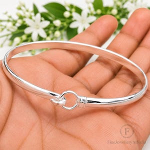 May include: A polished silver bangle bracelet featuring a smooth, rounded band and a circular hook-and-eye clasp mechanism. The metal surface is highly reflective, showcasing a clean, continuous design that connects at the front with a small ring.