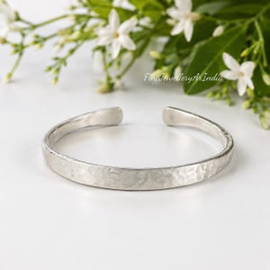 May include: A silver-toned cuff bracelet with a textured, hammered finish. The bracelet features an open-ended design with rounded, smooth terminals. The surface of the metal displays a series of small, irregular indentations across the band.