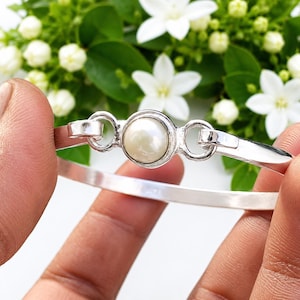 May include: A silver bangle bracelet featuring a single round white pearl set in a circular bezel at the center. The bracelet has a smooth, polished metallic finish and a simple, clean design with two circular links connecting the pearl setting.
