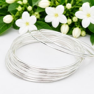 Op de afbeelding: Een set van meerdere dunne, gepolijste zilverkleurige armbanden die op elkaar zijn gestapeld. De cirkelvormige metalen banden hebben een glad, reflecterend oppervlak en een eenvoudig, onversierd ontwerp. De armbanden liggen in een licht overlappende groep.
