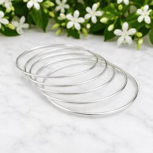 May include: A set of five thin, polished silver-tone bangle bracelets. Each bracelet is a simple, smooth, circular band with a reflective metallic finish. The bangles are stacked slightly overlapping each other, showcasing their uniform, slender design.