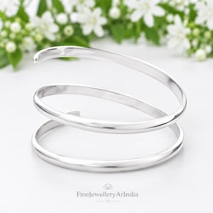 May include: A polished silver-toned cuff bracelet featuring a continuous spiral design with two overlapping loops. The metal surface is smooth and reflective, forming a circular shape that tapers at the ends. The text FineJewelleryArIndia is visible.