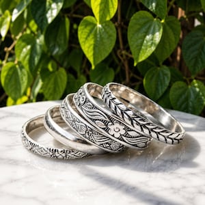 May include: A set of five silver-toned metal bangles stacked together. Each bracelet features distinct embossed patterns, including floral motifs, leaf-like textures, and geometric diamond shapes, with a polished finish and darkened recessed details.