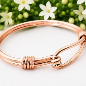 Może przedstawiać: Bransoletka typu bangle w kolorze różowego złota z zapięciem w stylu haczyka. Bransoletka wykonana jest z dwóch równoległych metalowych drutów owiniętych razem na jednym końcu, z zakrzywionym mechanizmem haczykowym zabezpieczającym okrągłą obręcz.