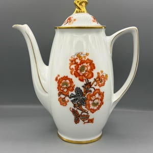 Op de afbeelding: Een witte porseleinen koffiepot met een bloemmotief van oranje en donkergrijze bloesems. De pot heeft een goudkleurige rand bij het handvat, de tuit, de voet en de bovenkant, en is voorzien van een bijpassend deksel met een gouden knop.