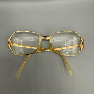May include: Gold-toned metal eyeglasses featuring rectangular lenses with rounded corners. The frame includes a central bridge with a small cutout detail and thin temples that extend outward, ending in clear, curved plastic tips for comfort.