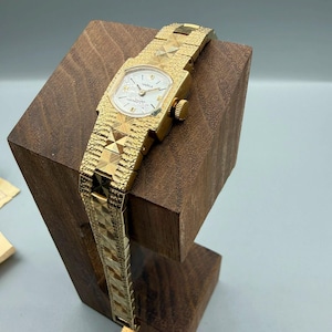 May include: A gold-tone wristwatch featuring a textured, geometric link bracelet and a rectangular case with a white dial. The watch face displays the text "ЧАЙКА" and "СДЕЛАНО В СССР" in black lettering, with gold-tone hands and hour markers.