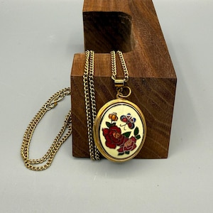 Pode incluir: Um pingente de medalhão oval em tom dourado numa corrente a condizer. O medalhão apresenta um embutido de esmalte em tom creme decorado com um gráfico colorido de duas rosas vermelhas, folhas verdes e uma pequena borboleta azul e laranja.