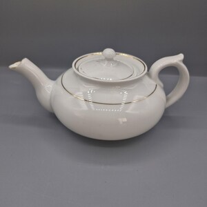May include: A white ceramic teapot with a rounded body, a curved handle, and a spout. The lid features a small knob handle. A thin, metallic gold band encircles the lid and the upper portion of the teapot body, providing a subtle decorative accent.