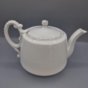May include: A white ceramic teapot featuring a smooth, rounded body, a matching lid with a small knob handle, and a curved spout. The handle is ornate with decorative scrollwork details. The rim of the lid and the spout tip have thin gold trim.
