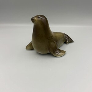 May include: A glossy ceramic figurine of a seal in a seated position. The seal features a smooth, gradient finish transitioning from a dark brown head to a lighter, muted olive-brown body, with detailed flippers and a small, dark snout.
