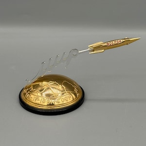May include: A gold-colored Vostok rocket model mounted on a clear acrylic stand shaped like cursive Cyrillic text. The rocket features red lettering. It sits on a gold dome base with a map engraving and the text 12-IV-1961-CCCP on a black rim.