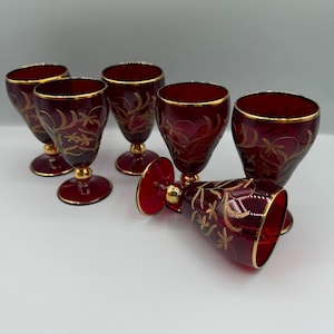 Peut inclure: Un ensemble de six verres à pied en verre rouge rubis avec des gravures florales dorées et des bords dorés. Chaque verre possède une tige courte avec un élément décoratif doré au-dessus d'une base circulaire. Un verre est couché.