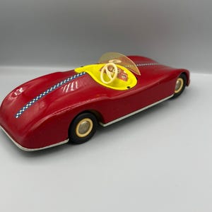 Pode incluir: Um carro de corrida de brinquedo vermelho vintage com cockpit amarelo, volante cor de creme e para-brisa translúcido. A carroceria apresenta uma faixa xadrez azul e branca no centro e rodas pretas com aros na cor creme.