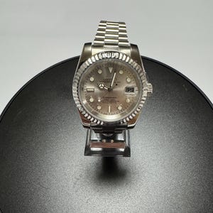 Peut inclure: Une montre-bracelet argentée avec un bracelet à maillons métalliques, une lunette cannelée et un cadran circulaire doté d'index lumineux, d'aiguilles argentées, d'un guichet de date à trois heures et du texte Seiko Oyster Perpetual Submariner.