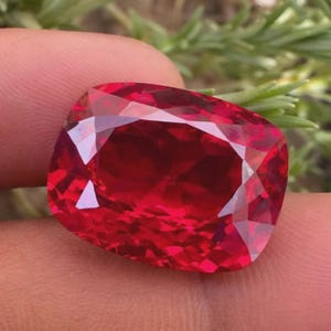 May include: A cushion-cut gemstone with a deep, vibrant red colour and a faceted surface that reflects light. The stone features a rectangular shape with rounded corners and a high-clarity, transparent appearance, showcasing internal light refraction.
