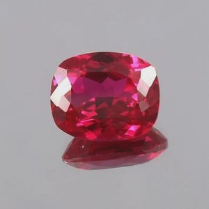 May include: A vibrant, deep red gemstone with a cushion cut, featuring multiple triangular and trapezoidal facets that create a brilliant, reflective surface. The stone displays a rich, saturated crimson hue with clear light refraction.