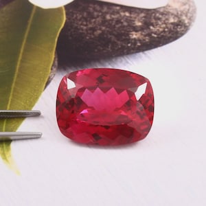 May include: A cushion-cut, deep red gemstone with a faceted surface that reflects light. The stone is positioned next to a pair of silver-toned metal tweezers, showcasing its vibrant colour and precise geometric cut in a clear, focused display.