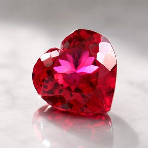 Puede incluir: Una piedra preciosa en forma de corazón de color rojo intenso y vibrante con una superficie facetada. La piedra presenta un corte geométrico complejo que crea múltiples planos que reflejan la luz, resultando en un aspecto brillante.