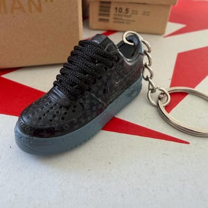 Può includere: Un portachiavi a forma di sneaker in miniatura con un motivo a monogramma ripetuto sulla tomaia, lacci neri e una suola grigio scuro. Il lato della scarpa mostra la scritta AIR e un anello portachiavi in metallo argentato è fissato al tallone.