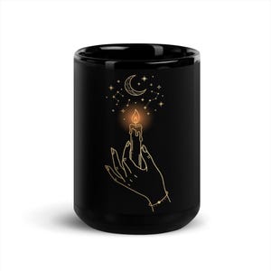May include: A black ceramic mug featuring a gold-toned line art illustration of a hand holding a lit candle. Above the candle flame, there is a crescent moon surrounded by small stars and constellations, all rendered in a delicate gold-colored outline.