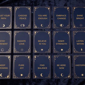 May include: A set of fifteen rectangular cards with a dark blue background and gold foil accents. Each card features a unique inspirational phrase such as Trust Your Path, Choose Peace, You Are Enough, and Unstoppable, framed by celestial motifs.