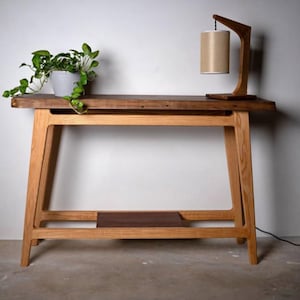Pode incluir: Uma mesa consola com um tampo de madeira castanho-escuro de bordo natural e uma estrutura de madeira clara com uma prateleira inferior. Uma planta verde num vaso cinzento e um candeeiro de mesa de madeira com cúpula cilíndrica bege estão em cima.
