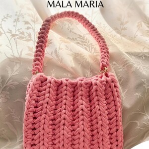 Pode incluir: Uma bolsa de crochê rosa com uma alça trançada grossa e textura canelada vertical. O texto TUTORIAL PASSO A PASSO MALA MARIA é exibido no topo, e um pequeno gráfico de abelha com o texto ABELINHA e por @abelinha.croche está na parte inferior.