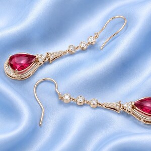 May include: A pair of drop earrings crafted from rose gold, each featuring a prominent pear-shaped red gemstone at the base. The design includes three small round clear stones along the hook and a decorative setting accented with tiny white diamonds.