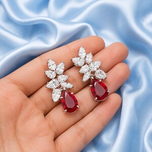 May include: A pair of silver-toned earrings featuring a cluster of clear, marquise-cut stones arranged in a leaf-like pattern, with a single, large, pear-shaped deep red stone dangling from the bottom of each earring setting.