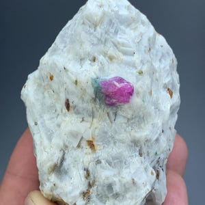 Könnte beinhalten: Ein rohes, kristallines Mineralexemplar mit einer großen, undurchsichtigen, quarzähnlichen weißen Matrix, einem eingebetteten, leuchtend pinkfarbenen Rubinkristall und einem kleinen Fleck aus blaugrünem Mineral auf der rauen Oberfläche.
