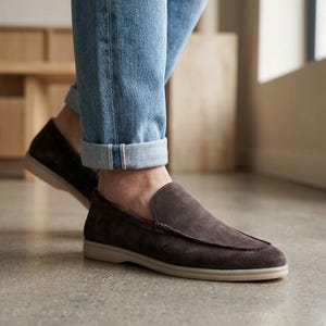 May include: A pair of dark brown suede loafers featuring a moccasin-style stitched toe seam and a flat, light-colored rubber sole. The shoes are worn with cuffed light blue denim jeans that reveal the ankle area above the shoe opening.