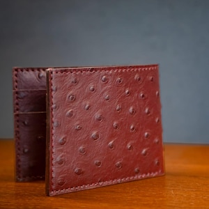 Puede incluir: Una cartera bifold de cuero color burdeos profundo con un patrón distintivo de plumas de avestruz con protuberancias en relieve en la superficie. La cartera muestra costuras visibles a lo largo de los bordes y varias ranuras interiores para tarjetas.