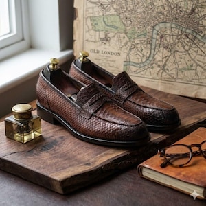 May include: A pair of brown loafers with a textured snakeskin pattern, featuring a classic penny slot strap across the vamp, a rounded toe, and a low stacked heel, displayed with gold-toned shoe trees inserted inside each shoe.