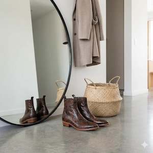 Può includere: Un paio di stivaletti in pelle marrone con motivo a pelle di serpente e tacchi bassi, posizionati su un pavimento in cemento accanto a un grande specchio rotondo, un cesto in vimini intrecciato e un cappotto di lana beige appeso.